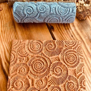 Puede incluir: Un rodillo estampado azul claro y un sello rectangular de color terracota, ambos con un diseño en espiral. El rodillo es cilíndrico, mientras que el sello es plano. El diseño recuerda a fósiles o conchas.