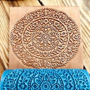 May include: A bright blue cylindrical rolling pin with a raised floral mandala pattern, positioned next to a square piece of tan clay that displays the same intricate, embossed circular design pressed into its surface with clear detail.
