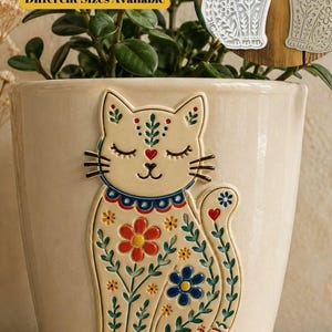 May include: A cream-coloured ceramic planter featuring a raised, hand-painted cat design with floral patterns in red, blue, and green. The cat has closed eyes and a heart-shaped nose. Two white cat-shaped clay cutters are visible in the upper right.