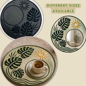 May include: Three circular ceramic items featuring embossed monstera leaves, a sun, and wavy lines. One is a dark grey wall plaque, one is a mirror with a green and yellow finish, and one is a beige serving tray holding a matching coffee cup and saucer.
