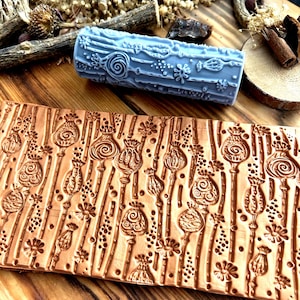 Op de afbeelding: Een lichtblauwe, cilindrische embossing roller met bloemen- en wervelontwerpen, en een bruine leren embossing sheet met een klaproos- en bloemenpatroon. De items liggen op een houten oppervlak.
