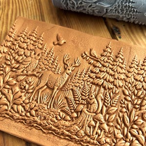 May include: A detailed, brown, embossed leather craft depicting a forest scene with a deer, squirrel, birds, and trees. A grey roller with a forest pattern is in the background. The craft is likely for decorative or crafting purposes.