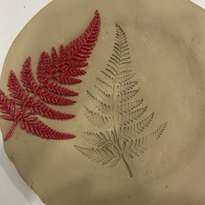 May include: A round, beige clay plate with two fern leaf impressions. One is a raised, red, glittery fern, and the other is an indented fern. The plate is a natural, earthy tone, perfect for craft projects or home decor.