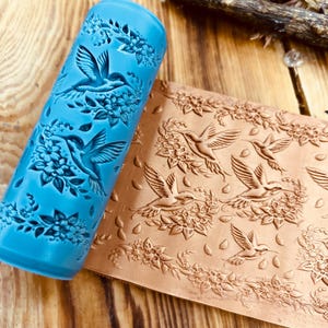 May include: A blue rolling pin with a hummingbird and floral design next to a brown embossed sheet with the same pattern. The rolling pin is cylindrical and features the design carved into its surface. The embossed sheet shows the pattern in detail.