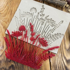 May include: An off-white rectangular mould with an embossed floral design, showcasing various plant silhouettes. A separate, vibrant red, glittery floral piece is placed in front. The image is set on a wooden surface.