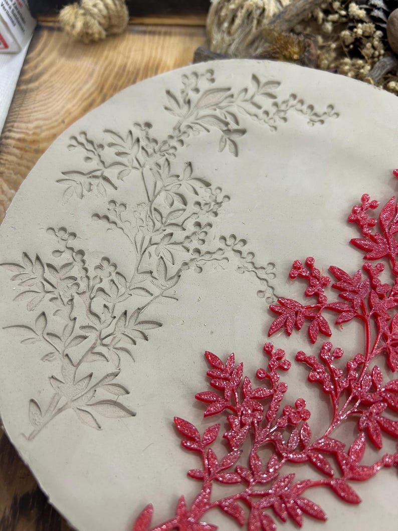 May include: A round, off-white mold with a floral design carved into the surface. A sprig of red, glittery leaves and berries is placed on the right side of the mold, creating a contrast in color and texture. The mold is likely used for crafting.