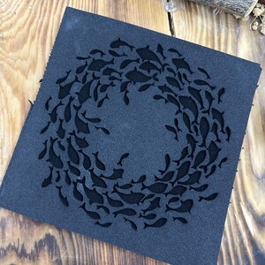 May include: A square, dark gray leather coaster featuring a school of fish swimming in a circular pattern. The fish are cut out of the leather, creating a negative space design. The coaster is placed on a wooden surface.