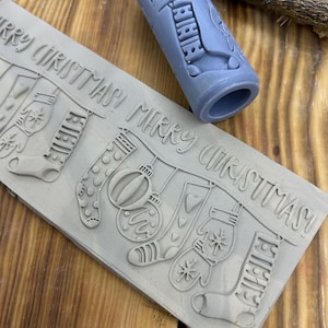 Puede incluir: Un sello de goma gris con las palabras "MERRY CHRISTMAS" e imágenes festivas navideñas. El sello incluye calcetines, adornos y mitones. Un sello cilíndrico con la palabra "12AM" está al lado del sello rectangular.