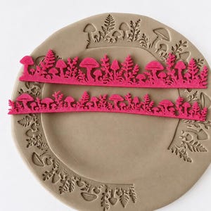 May include: Two long, thin, bright pink strips featuring a detailed cutout pattern of various mushrooms and fern-like foliage. These strips are positioned horizontally across a circular, light brown clay plate that has matching mushroom and fern impressions.