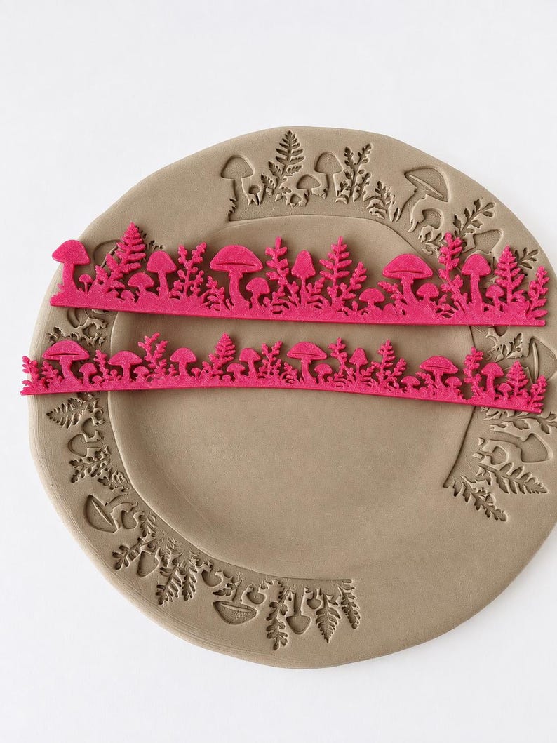 May include: Two long, thin, bright pink strips featuring a detailed cutout pattern of various mushrooms and fern-like foliage. These strips are positioned horizontally across a circular, light brown clay plate that has matching mushroom and fern impressions.