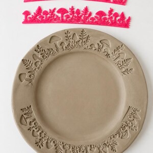 May include: Two bright pink flexible silicone stamps featuring intricate mushroom and fern patterns, positioned above a circular gray clay plate that displays the same embossed botanical designs pressed into its outer rim surface.