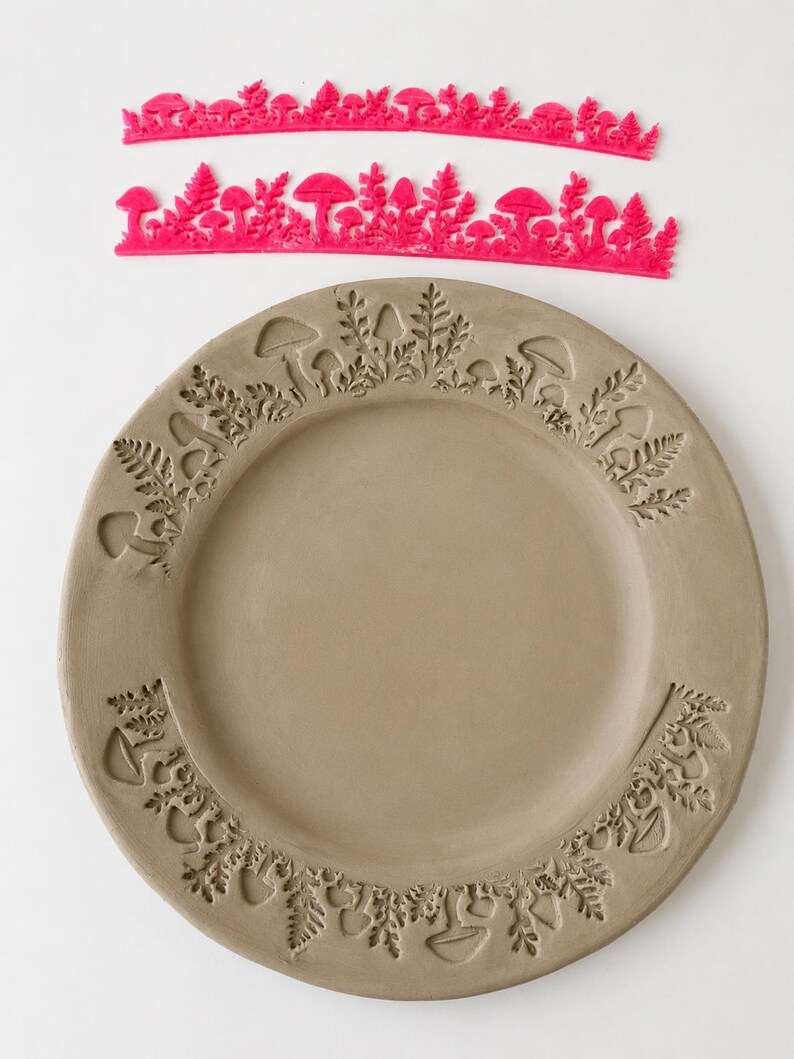 May include: Two bright pink flexible silicone stamps featuring intricate mushroom and fern patterns, positioned above a circular gray clay plate that displays the same embossed botanical designs pressed into its outer rim surface.