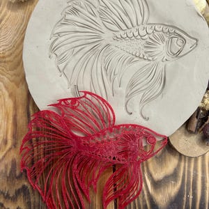 May include: A red, laser-cut betta fish sits below a circular, light-colored clay impression of a betta fish. The clay impression has detailed lines. The fish has flowing fins and tail.