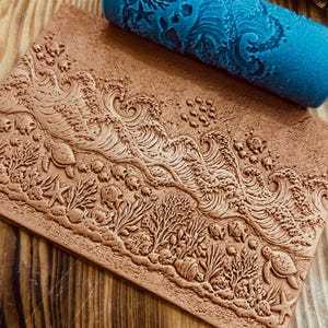 May include: A brown clay slab and a blue roller with an ocean-themed design. The slab features detailed relief carvings of waves, coral, turtles, and starfish. The blue roller has a matching ocean pattern, suggesting a tool for imprinting designs.