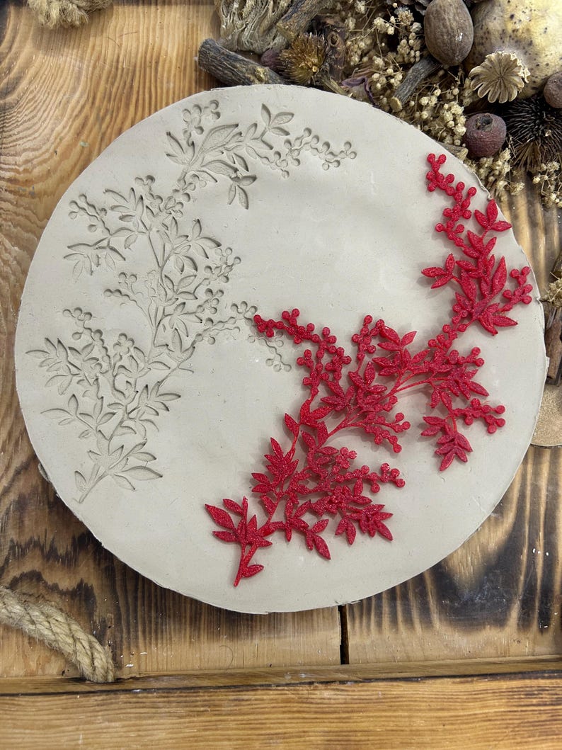 May include: A round, off-white decorative piece with floral designs. One side features an embossed floral pattern, while the other side has a vibrant red, three-dimensional floral embellishment. The piece is set against a rustic wooden background.