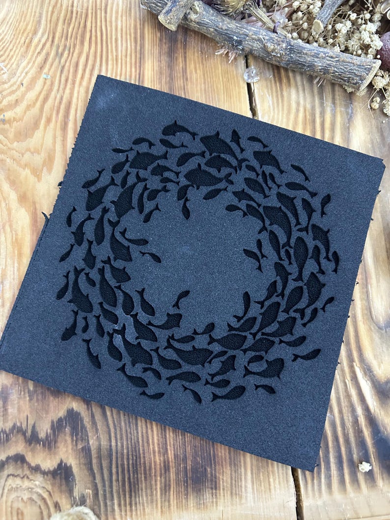 May include: A square, dark gray leather coaster featuring a school of fish swimming in a circular pattern. The fish are cut out of the leather, creating a negative space design. The coaster is placed on a wooden surface.