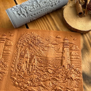 May include: A detailed, gray roller with a nautical design sits above a terracotta-colored, embossed piece. The embossed piece features a lighthouse, a house, and ocean waves. The scene is reminiscent of a coastal landscape.