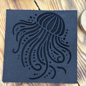 Puede incluir: Un posavasos cuadrado negro con un diseño detallado de medusa. La medusa presenta tentáculos fluidos y un cuerpo texturizado, creando un contraste con la superficie lisa del posavasos. Fabricado con un material duradero.