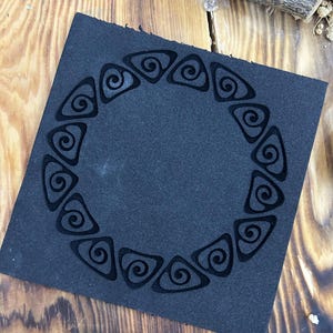 Puede incluir: Un posavasos cuadrado de cuero negro con un diseño decorativo de espirales repetidas en forma circular. El posavasos está colocado sobre una superficie de madera con vetas naturales.
