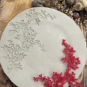 May include: A round, light gray ceramic plate with an embossed floral design. A red, glittery floral accent is placed on the plate. The plate is displayed on a wooden surface with dried flowers and twine.