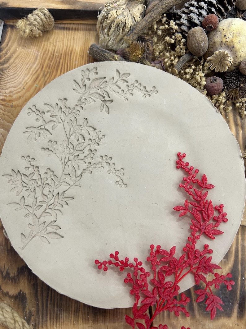 May include: A round, light gray ceramic plate with an embossed floral design. A red, glittery floral accent is placed on the plate. The plate is displayed on a wooden surface with dried flowers and twine.