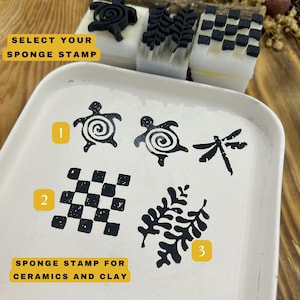 May include: A white ceramic tray with black sponge stamps for ceramics and clay. The tray features stamped designs of turtles, a checkerboard, and leaves. The text "SELECT YOUR SPONGE STAMP" and "SPONGE STAMP FOR CERAMICS AND CLAY" are visible.
