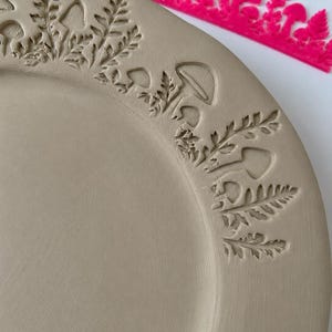 May include: A light beige ceramic plate featuring a stamped border design of mushrooms and fern leaves. A bright pink plastic stamp tool with a matching mushroom and fern pattern is positioned along the top edge of the plate.