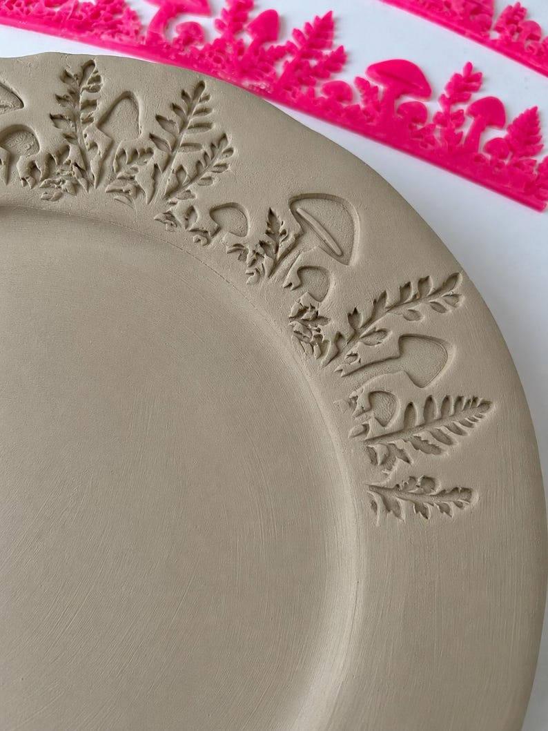May include: A light beige ceramic plate featuring a stamped border design of mushrooms and fern leaves. A bright pink plastic stamp tool with a matching mushroom and fern pattern is positioned along the top edge of the plate.
