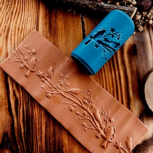 May include: A terracotta-colored rectangular stamp with an embossed design of birds on flowering branches, next to a blue cylindrical stamp with a matching design. The stamps are on a wooden surface.