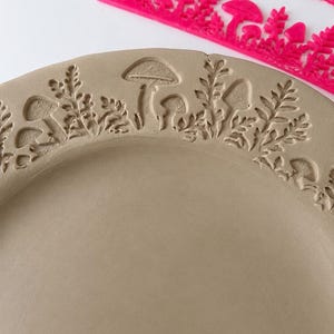 May include: A light brown circular clay plate featuring an embossed pattern of mushrooms and ferns along the rim. A bright pink plastic stamp tool with a matching mushroom and fern design is positioned above the plate on a white surface.