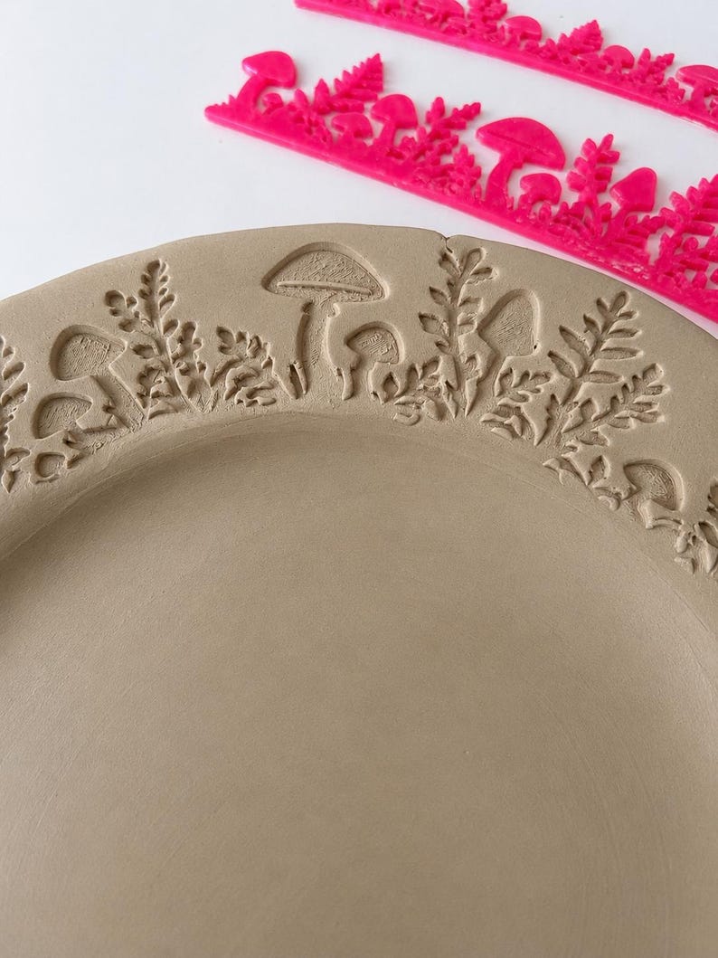 May include: A light brown circular clay plate featuring an embossed pattern of mushrooms and ferns along the rim. A bright pink plastic stamp tool with a matching mushroom and fern design is positioned above the plate on a white surface.