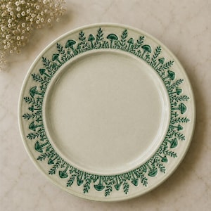 May include: A round ceramic plate with a cream-colored base and a raised, embossed border featuring a repeating pattern of dark green mushrooms and fern-like foliage. The center of the plate is smooth and plain, surrounded by the detailed relief design.