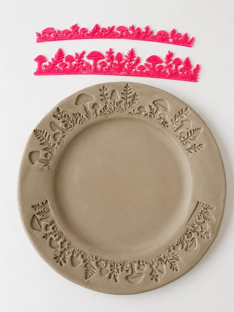 May include: Two bright pink flexible silicone stamps featuring intricate mushroom and fern patterns, positioned above a circular gray clay plate that displays the same embossed botanical designs pressed into its outer rim surface.