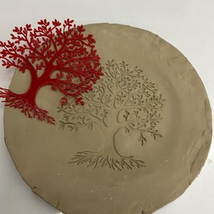 Könnte beinhalten: Ein rundes, hellbraunes Tonstück mit einem eingepressten Baumdesign. Ein roter, lasergeschnittener Baum liegt auf dem Ton. Das Baumdesign ist eine detaillierte Illustration eines Baumes mit Wurzeln, Stamm und Ästen.