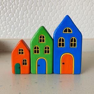 May include: Three colorful wooden houses in a row. The houses are painted in bright colors: orange, green, and blue. Each house has yellow-framed windows and a contrasting door. The houses are of varying heights, creating a playful arrangement.