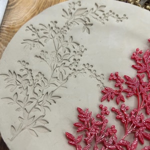 May include: A round, off-white mold with a floral design. The mold has an embossed pattern of leaves and berries. A separate, red, glittery floral piece is placed on the mold, showcasing the potential finished product.