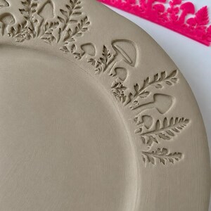May include: A light beige ceramic plate featuring a stamped border design of mushrooms and fern leaves. A bright pink plastic stamp tool with a matching mushroom and fern pattern is positioned along the top edge of the plate.