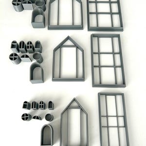 May include: A collection of gray plastic cookie cutters in various shapes, including arched windows, house frames, and rectangular window frames. There are also smaller shapes like circles and crosses. The cutters are arranged on a white surface.