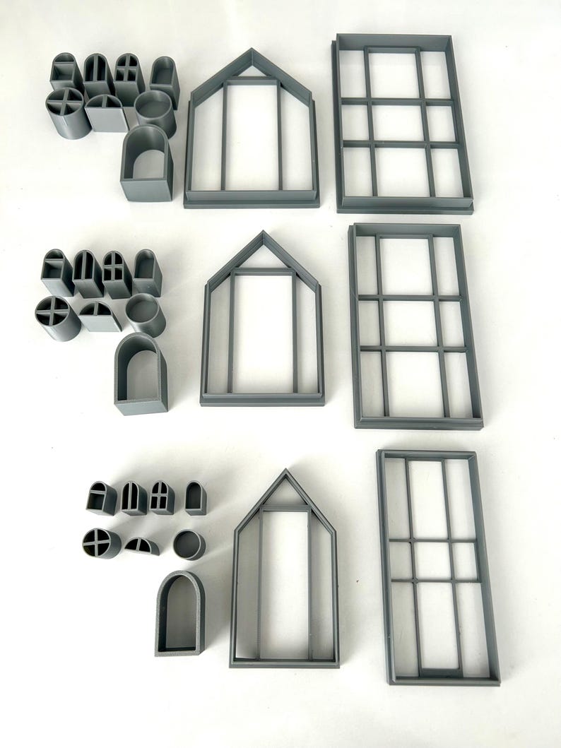 May include: A collection of gray plastic cookie cutters in various shapes, including arched windows, house frames, and rectangular window frames. There are also smaller shapes like circles and crosses. The cutters are arranged on a white surface.