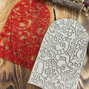 May include: Two decorative, arched-top stamps. One is red with a floral and bird design. The other is a light gray with an embossed bird and floral pattern. Both are on a wooden surface.