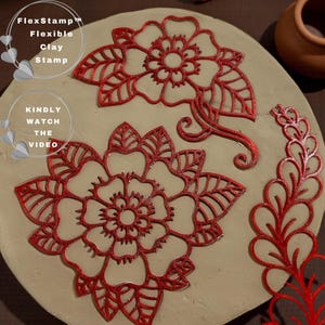 May include: A close-up of a clay stamp set with red floral designs. The set includes two large flower stamps, a decorative stem, and the text "FlexStamp Flexible Clay Stamp" and "KINDLY WATCH THE VIDEO". The stamps are on a light-colored surface.