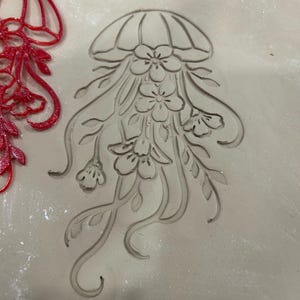 May include: A red jellyfish-shaped stamp with floral details is pressed into a light-colored clay surface. The stamp's design includes a rounded top, flowing tendrils, and flower accents, creating a decorative impression.