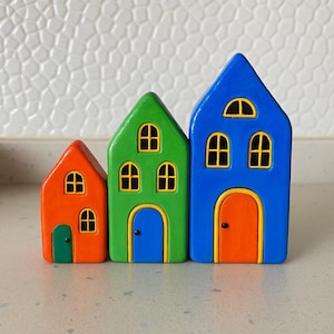 May include: Three colorful wooden houses in a row. The houses are painted orange, green, and blue, each with yellow-framed windows and a contrasting door. The houses are of varying heights, creating a playful, decorative display.