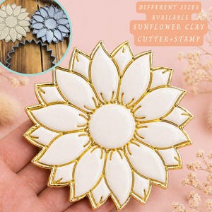 Op de afbeelding: Een witte en gouden zonnebloem klei uitsteker en stempel. De uitsteker heeft een gedetailleerd ontwerp met een wit centrum en bloemblaadjes, omlijnd in goud. De tekst "DIFFERENT SIZES AVAILABLE SUNFLOWER CLAY CUTTER+STAMP" is zichtbaar.