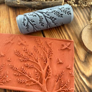 May include: A grey patterned roller and a terracotta-coloured stamp, both featuring leaf and tree designs. The roller is cylindrical, while the stamp is rectangular. These tools are likely for crafting or decorative use, with nature-inspired patterns.