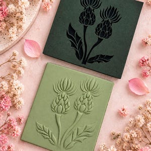 May include: Two rectangular tiles featuring a raised thistle plant design. The top tile is dark forest green with a black thistle motif, while the bottom tile is a lighter sage green with a matching embossed thistle pattern in the same colour.