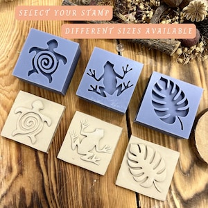 Può includere: Una collezione di timbri quadrati con vari disegni, tra cui una tartaruga, una rana e una foglia. I timbri sono nei colori beige chiaro e blu lavanda. Il testo "SELECT YOUR STAMP" e "DIFFERENT SIZES AVAILABLE" è visibile.