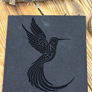 May include: A square, black coaster featuring a detailed, engraved hummingbird design. The bird is depicted in flight with outstretched wings and a long beak. The coaster is made of a dark, textured material.