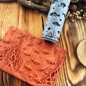 Può includere: Un mattarello azzurro con un design a tema oceanico con pesci, stelle marine e coralli. Un foglio di argilla color terracotta mostra una scena oceanica in rilievo abbinata. Gli oggetti sono su una superficie di legno.