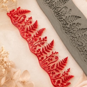 May include: A translucent red acrylic fern leaf stamp featuring intricate frond details alongside a rectangular gray clay slab embossed with the same matching fern leaf pattern, showcasing the texture transfer from the stamp to the clay material.